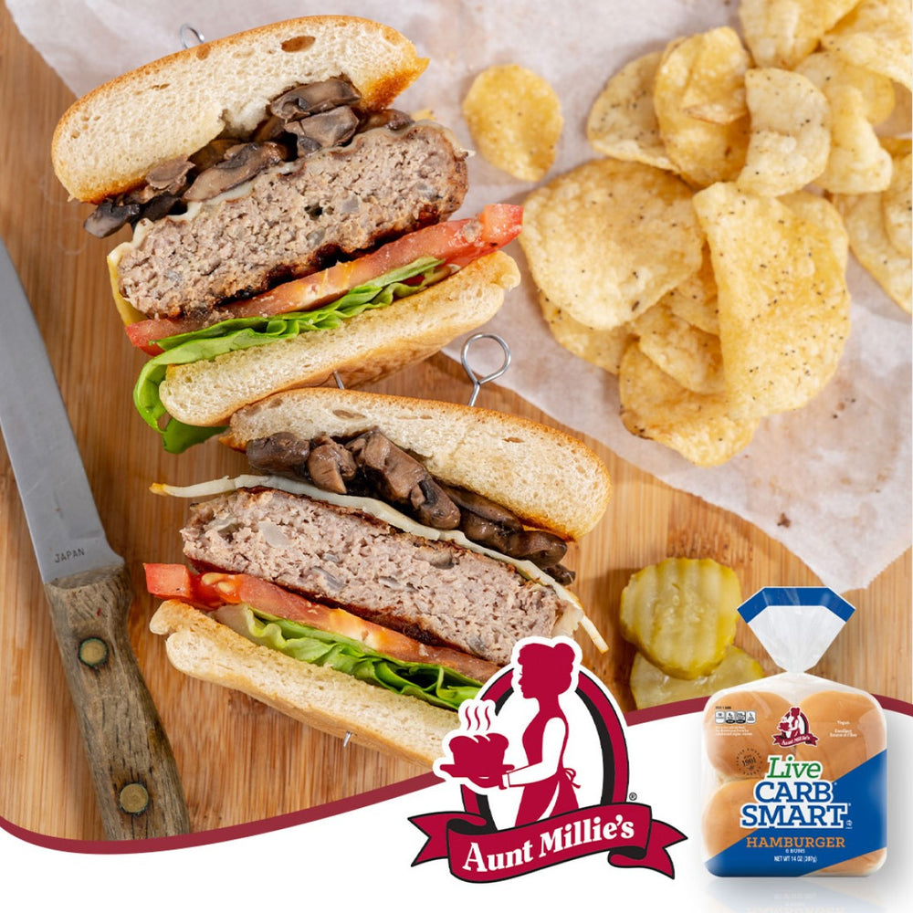A close-up of a sliced hamburger stacked with lettuce, tomato, and mushrooms, served with potato chips on a wooden board, alongside Aunt Millie's Carb-Smart hamburger buns and a knife.