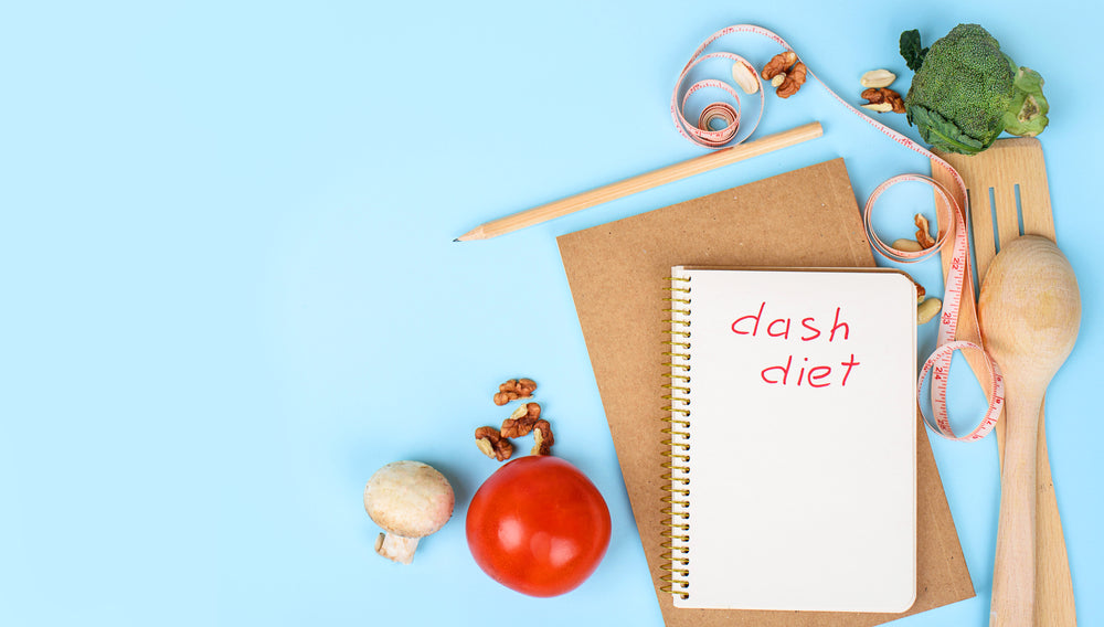 A notepad labeled "dash diet" sits on a light blue background surrounded by fresh vegetables, nuts, a pencil, measuring tape, and wooden utensils, suggesting a focus on healthy eating.