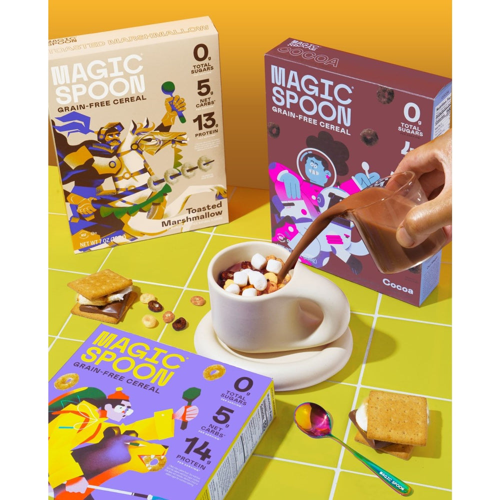 A hand pours chocolate milk into a white cup filled with cereal and marshmallows, surrounded by colorful boxes of Magic Spoon cereal and a graham cracker treat on a bright yellow table.