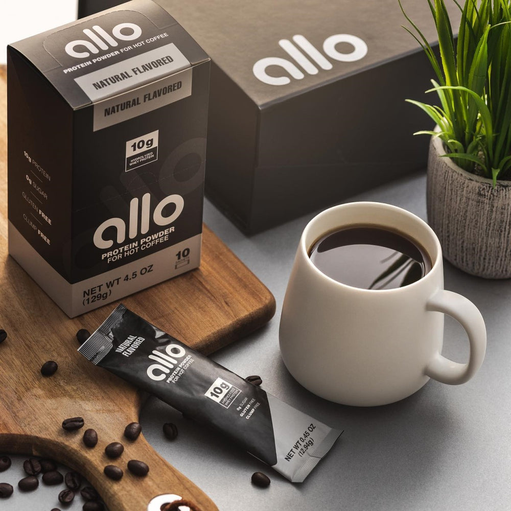 A white mug filled with hot coffee sits beside a packet of Allo protein powder and a box on a wooden cutting board, with coffee beans scattered around and a small potted plant in the background.