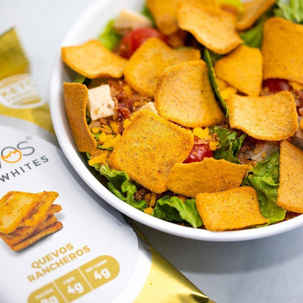 A bowl of salad features vibrant green lettuce, diced chicken, cherry tomatoes, shredded cheese, and topped with crispy, golden-brown snack squares, alongside a package of "Quevos Rancheros."