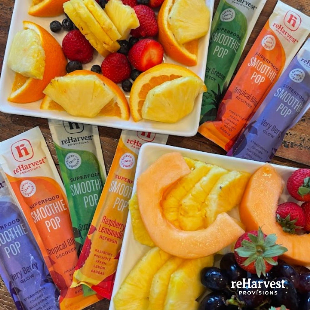 Colorful fruit slices, including oranges, strawberries, and pineapple, are arranged on two plates alongside several brightly packaged smoothie pops from reHarvest Provisions.