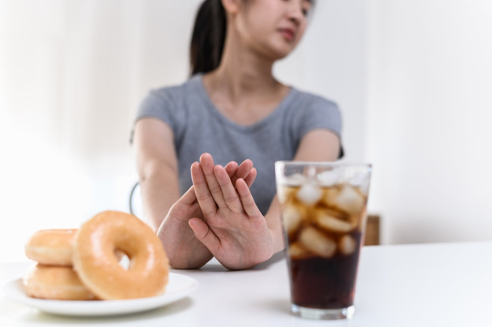 A young woman in a gray shirt sits at a table, holding her hand up in refusal while looking away from a plate of donuts and a glass of iced drink.