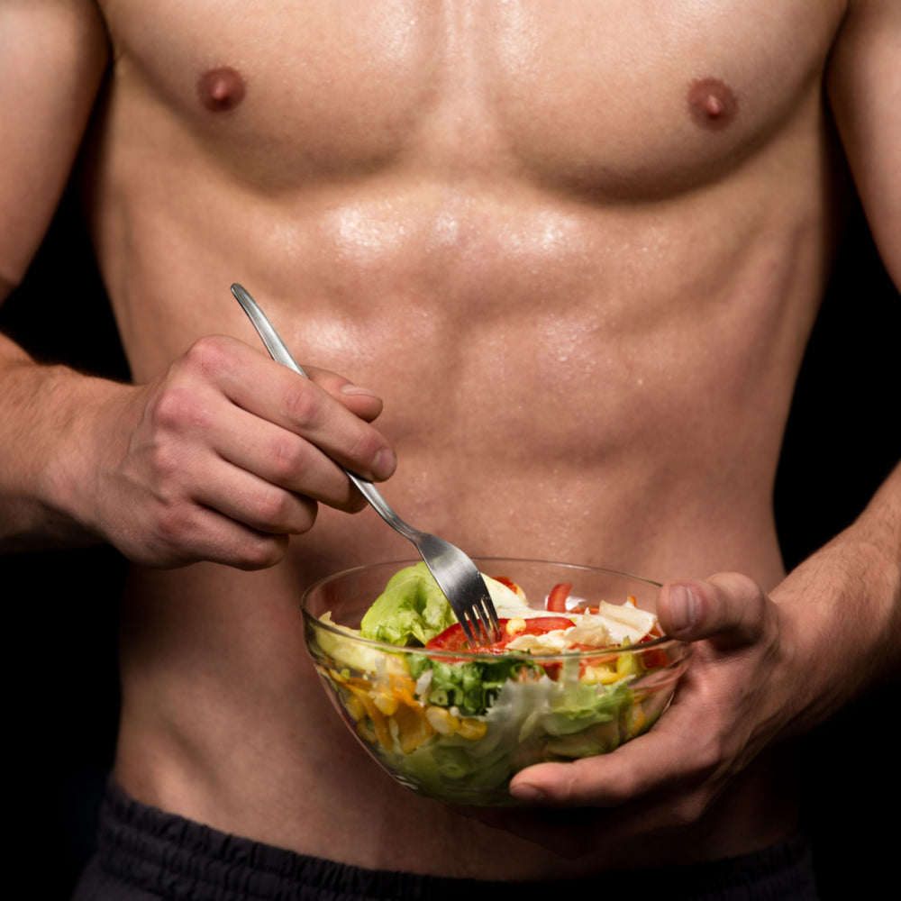 A muscular person holds a bowl of salad in one hand and a fork in the other, preparing to eat, with a dark background emphasizing their toned physique.