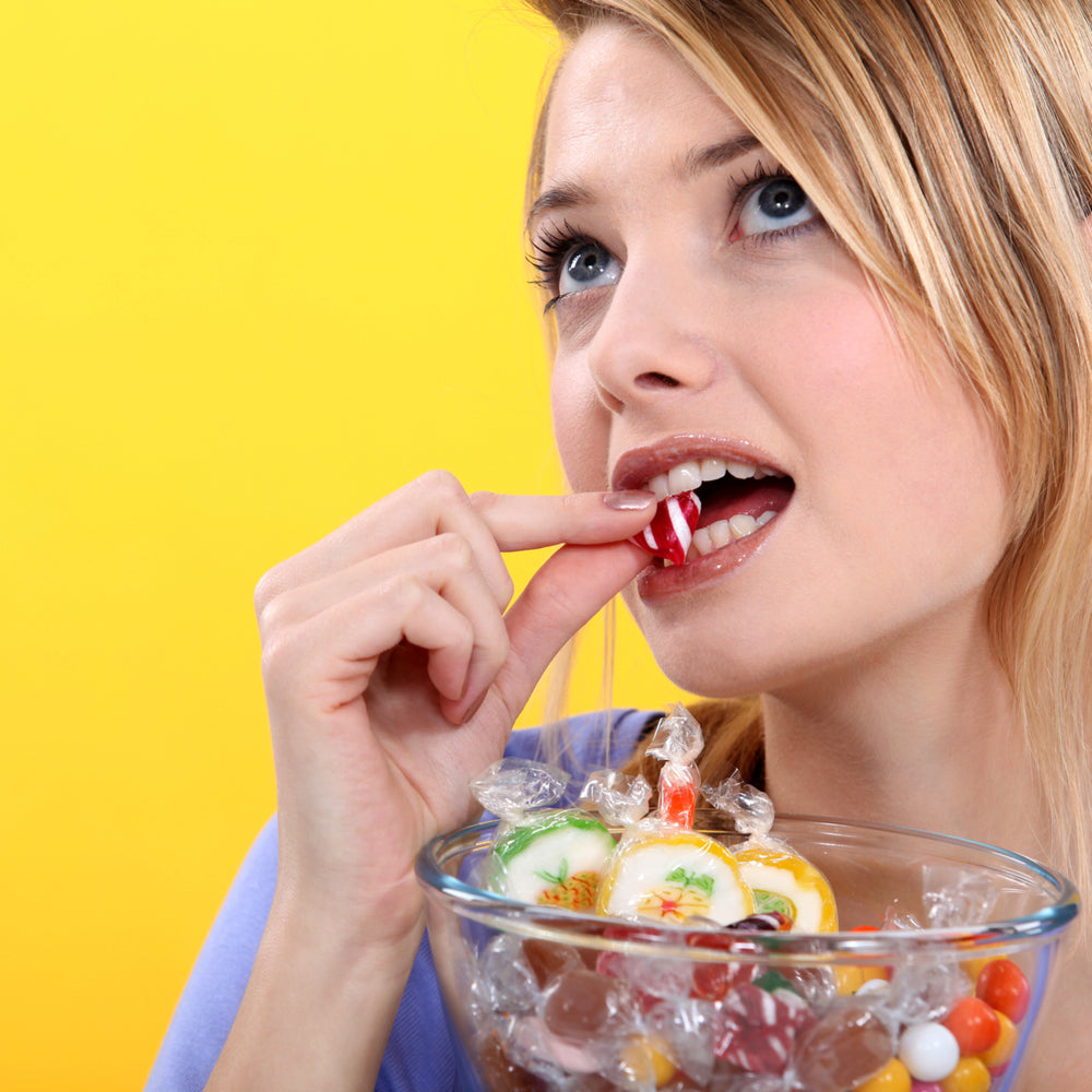 A young woman with blonde hair is placing a red and white striped candy into her mouth while looking up, with a bowl of assorted colorful candies in front of her against a bright yellow background.