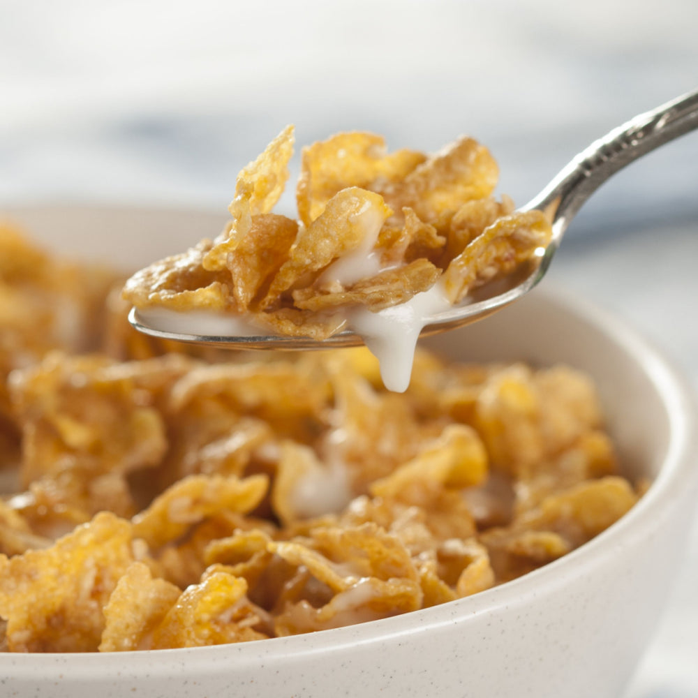 A spoonful of golden cornflakes topped with creamy milk is lifted from a bowl filled with more cornflakes, set against a softly blurred background.