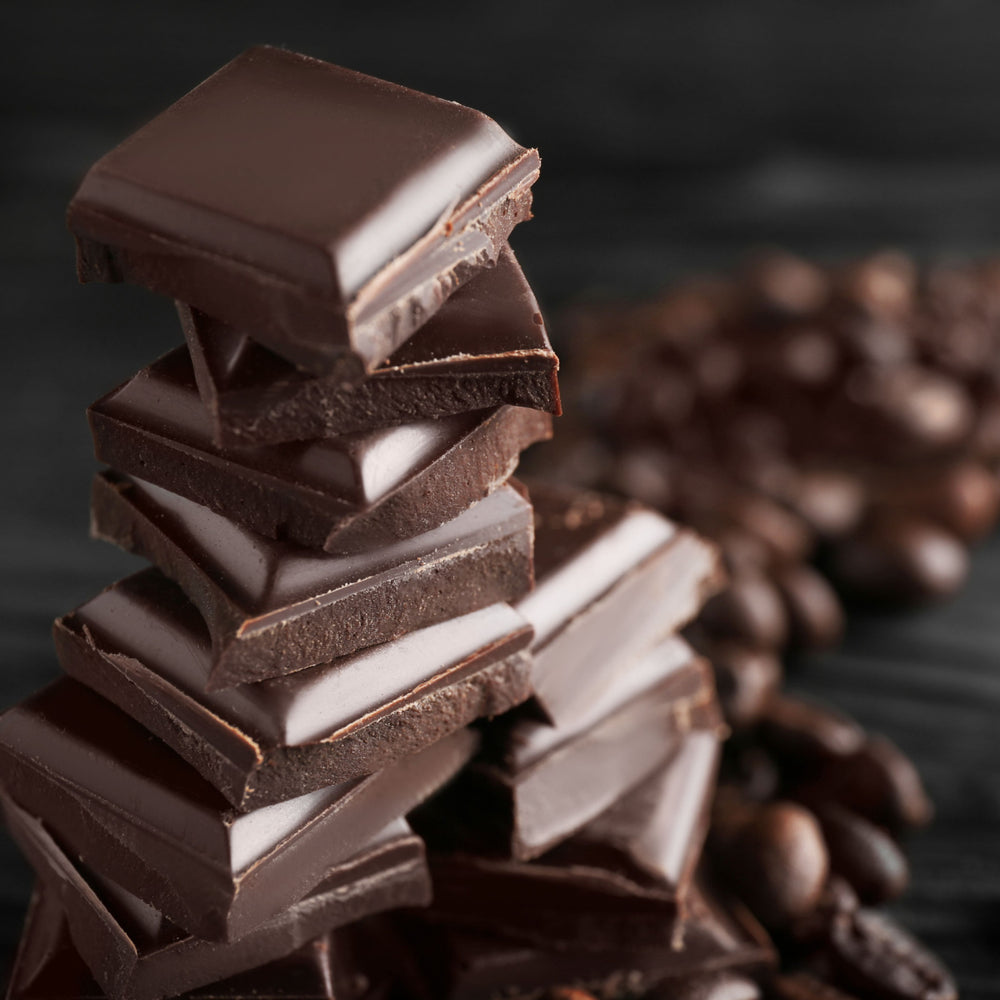 A stack of dark chocolate squares is arranged neatly on a dark surface, with roasted coffee beans scattered in the background, creating a rich and indulgent atmosphere.