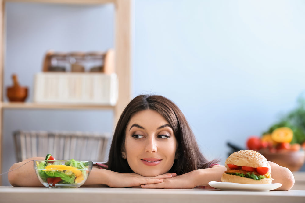 A woman with long brown hair rests her chin on her hands, looking thoughtfully at a hamburger and a bowl of salad on a table in a bright kitchen.