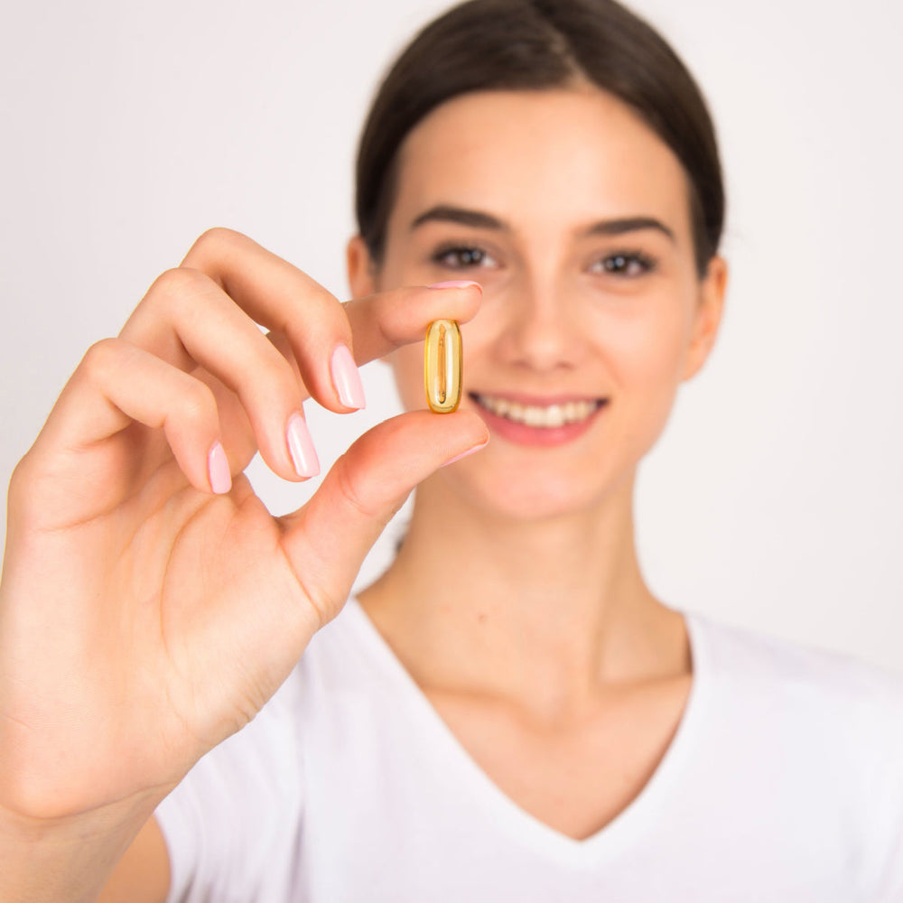 A smiling woman in a white shirt holds a golden capsule between her fingers against a plain background, suggesting a focus on health or wellness.