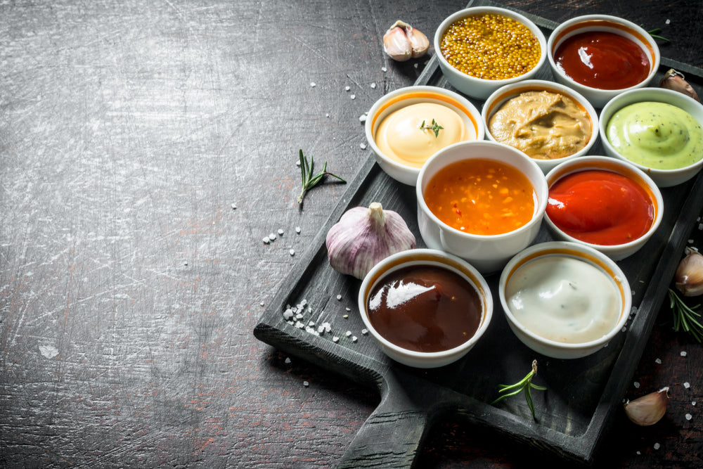 A wooden tray holds eight small bowls filled with various sauces, including mustard, ketchup, and a creamy dip, surrounded by garlic cloves and scattered salt on a dark surface.