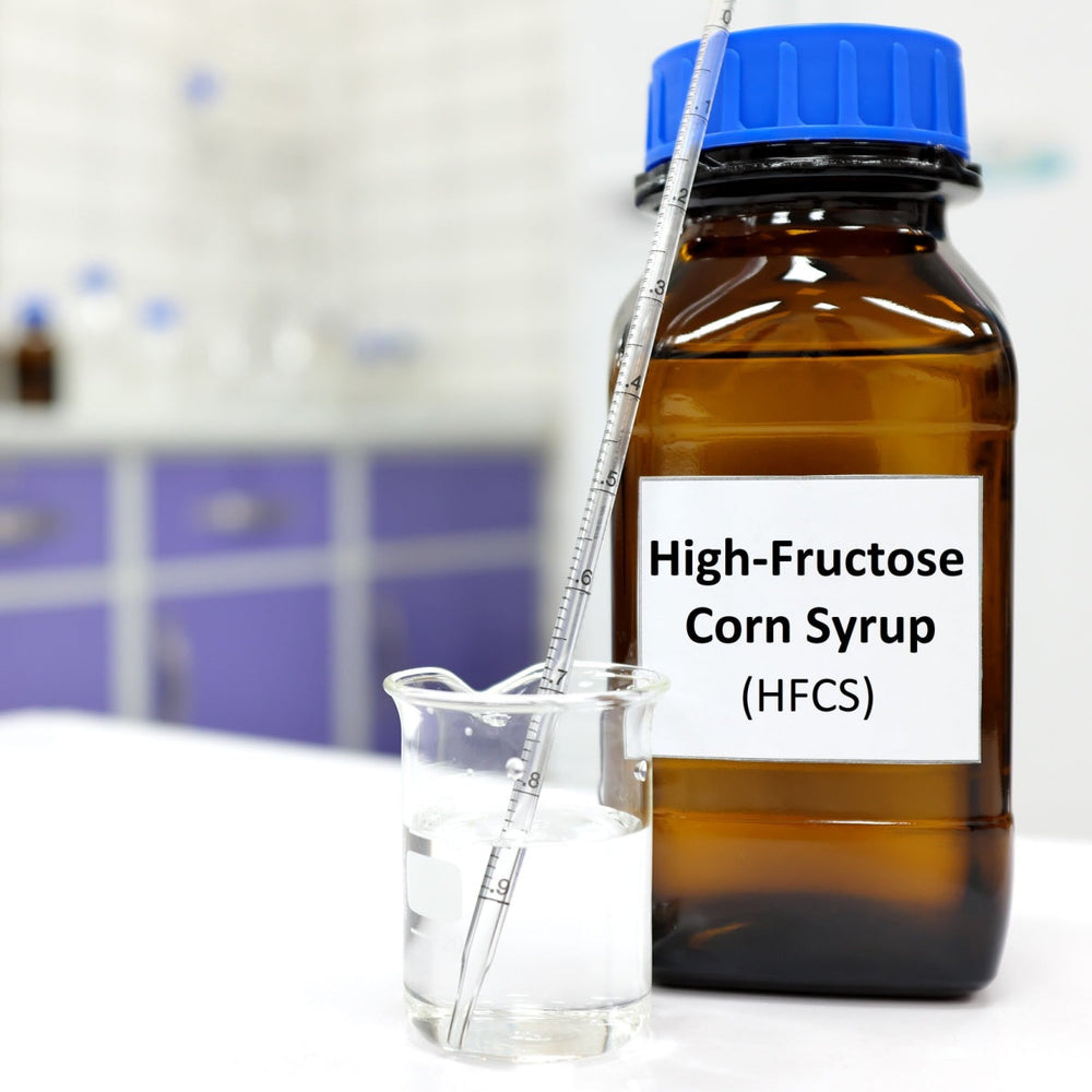 A brown bottle labeled "High-Fructose Corn Syrup (HFCS)" sits next to a clear beaker filled with liquid and a measuring stick, set against a blurred laboratory background.