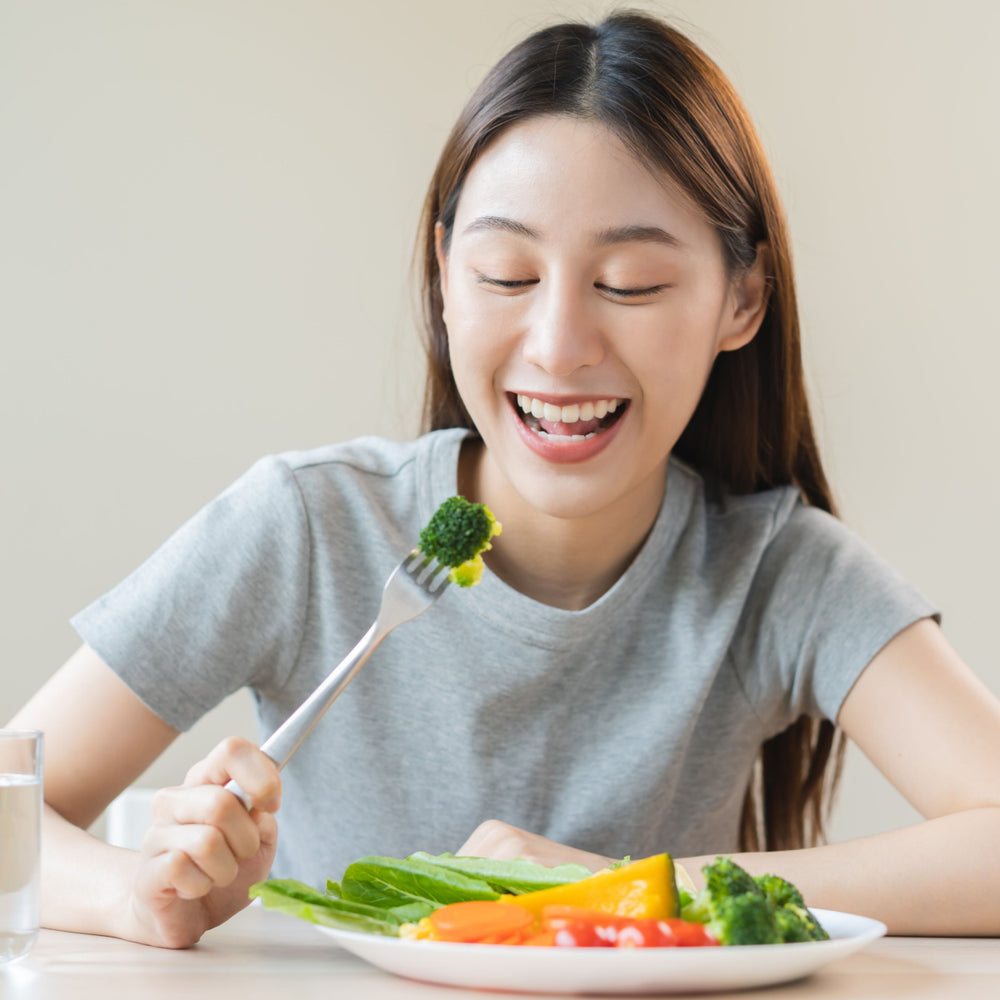 A young woman with long hair smiles joyfully while holding a fork with broccoli, sitting at a table with a colorful plate of vegetables and a glass of water beside her.