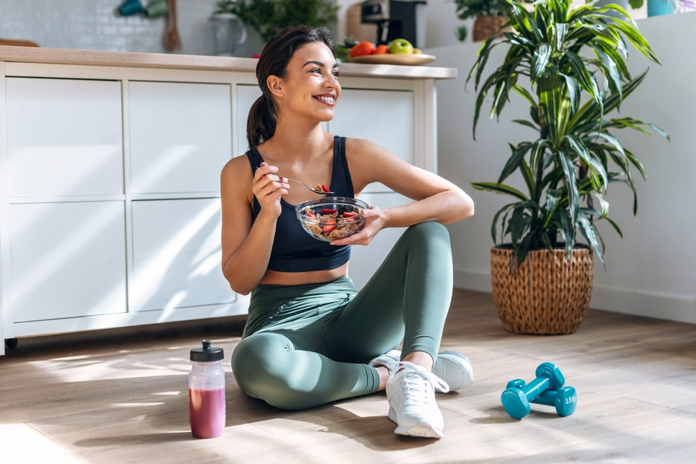 A woman sits on the floor in a bright kitchen, smiling while eating a bowl of fruit, with a water bottle and dumbbells nearby, surrounded by plants and healthy food on the counter.