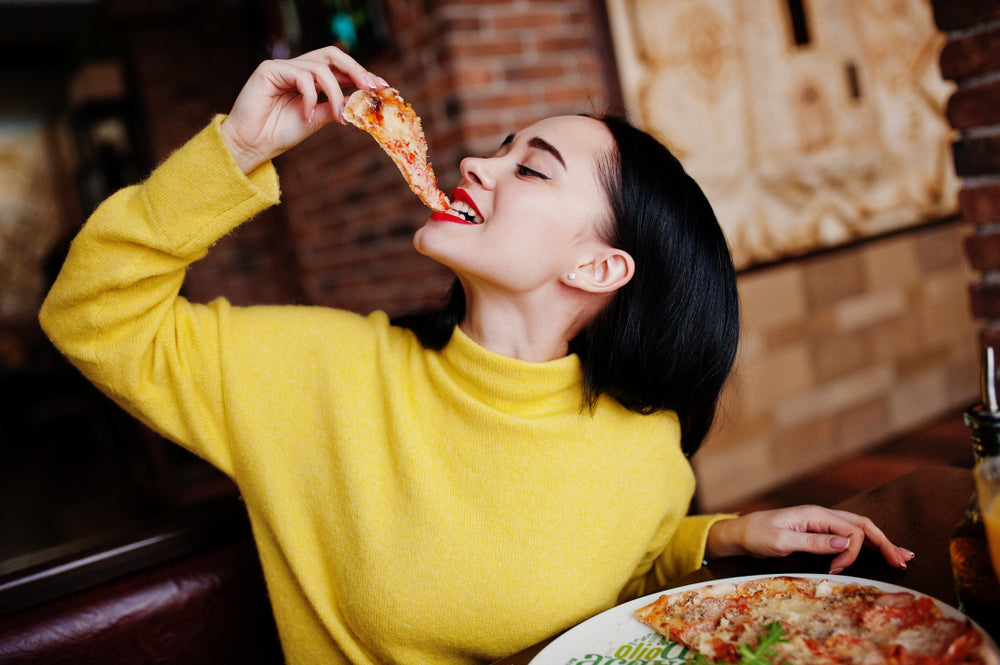 A woman in a bright yellow sweater joyfully takes a bite of pizza while seated at a table in a cozy restaurant with a brick wall in the background.