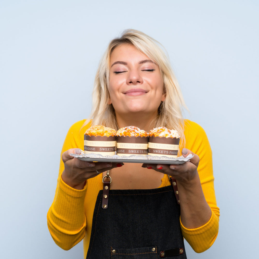 A smiling woman in a yellow sweater holds a plate with three decorated cupcakes, enjoying the moment against a light blue background.
