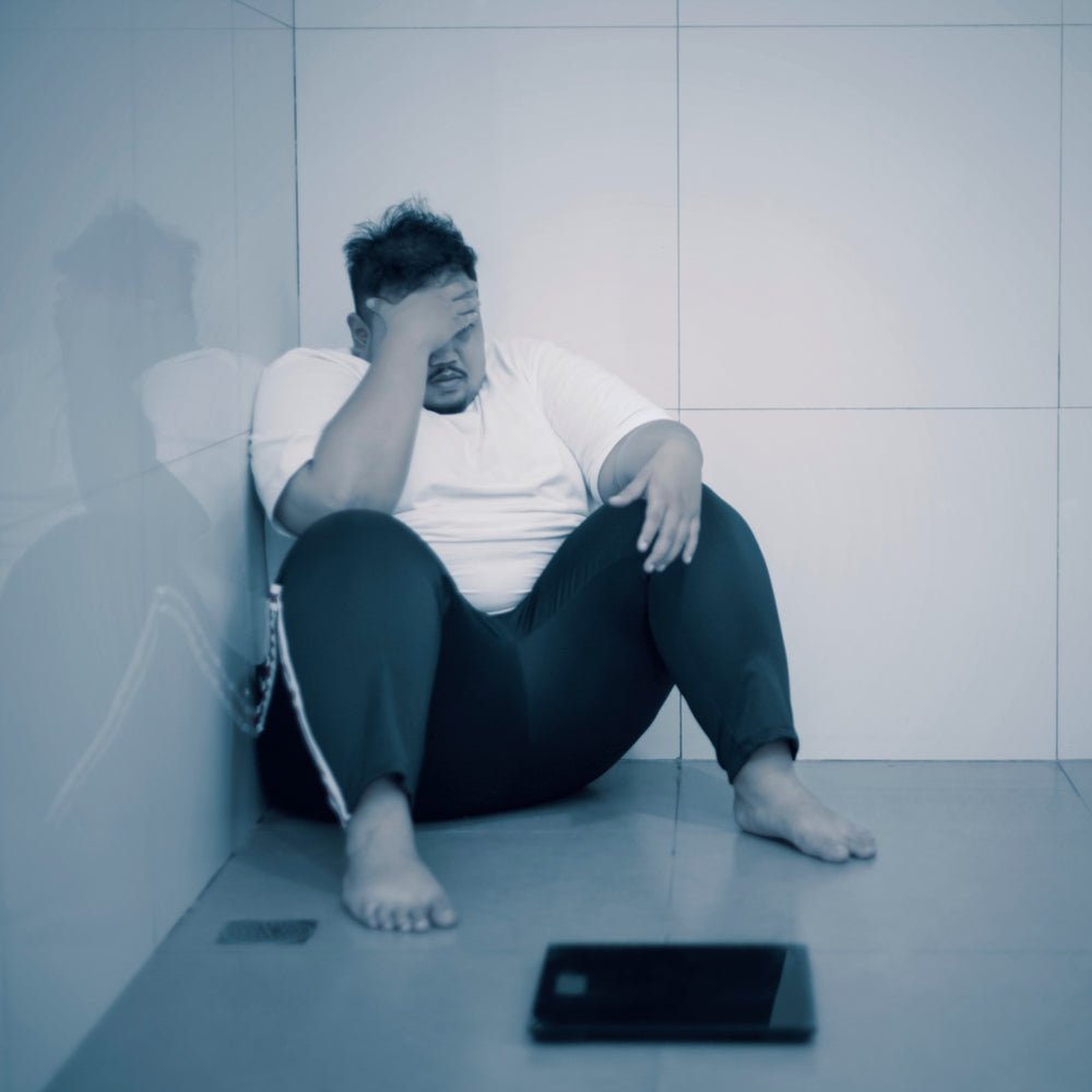 A person sits on the floor in a bathroom corner, holding their head in frustration, while a scale lies nearby on the tiled floor.