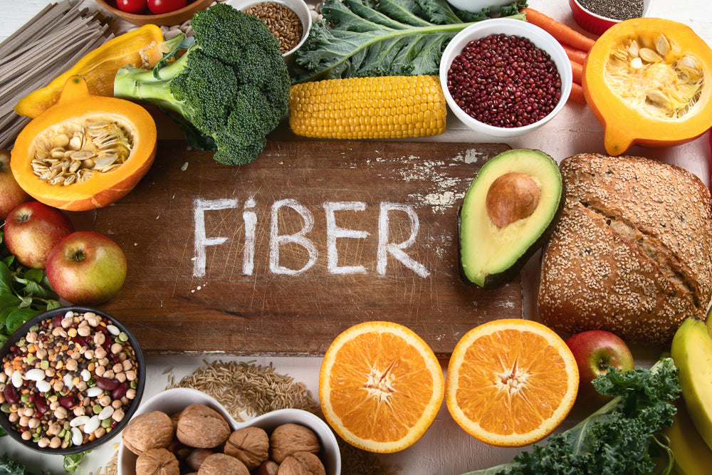 A wooden board displays the word "FIBER" surrounded by a variety of colorful fruits, vegetables, grains, and nuts, emphasizing healthy food choices rich in fiber.
