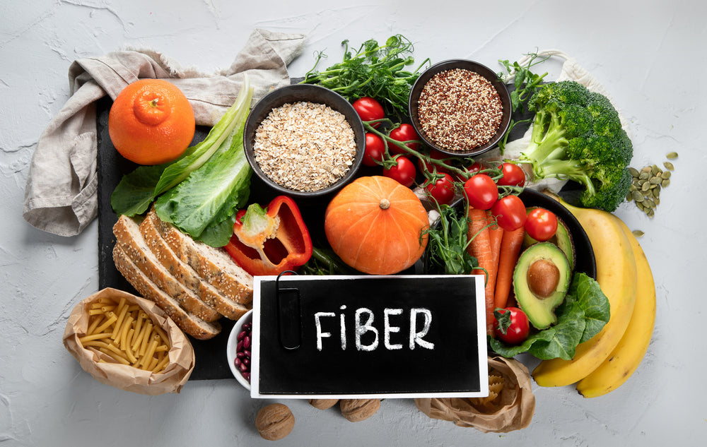 A variety of colorful fruits, vegetables, grains, and legumes are arranged on a table, with a chalkboard sign reading "FIBER" prominently displayed among them.