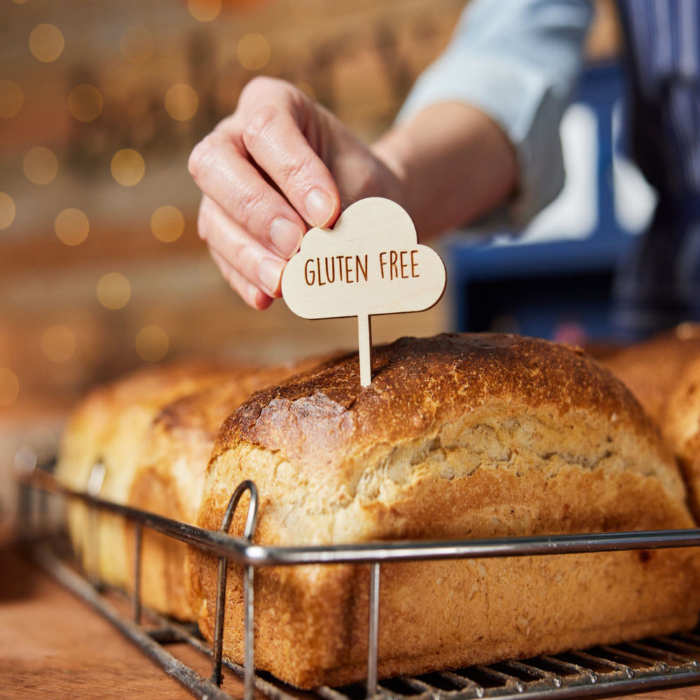 A hand places a wooden "Gluten Free" sign into a freshly baked loaf of bread, resting on a wire rack with a blurred, warm background of soft lights.