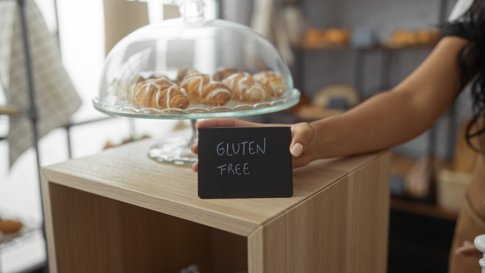 A person holds a small black sign that reads "GLUTEN FREE" in front of a display case filled with pastries, highlighting gluten-free options in a cozy, inviting setting.