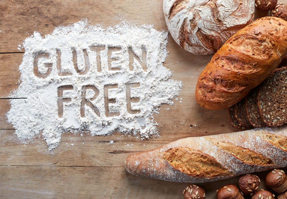 Flour is spread on a wooden surface with the words "GLUTEN FREE" written in it, surrounded by various types of bread, including a baguette and round loaves, emphasizing gluten-free baking.