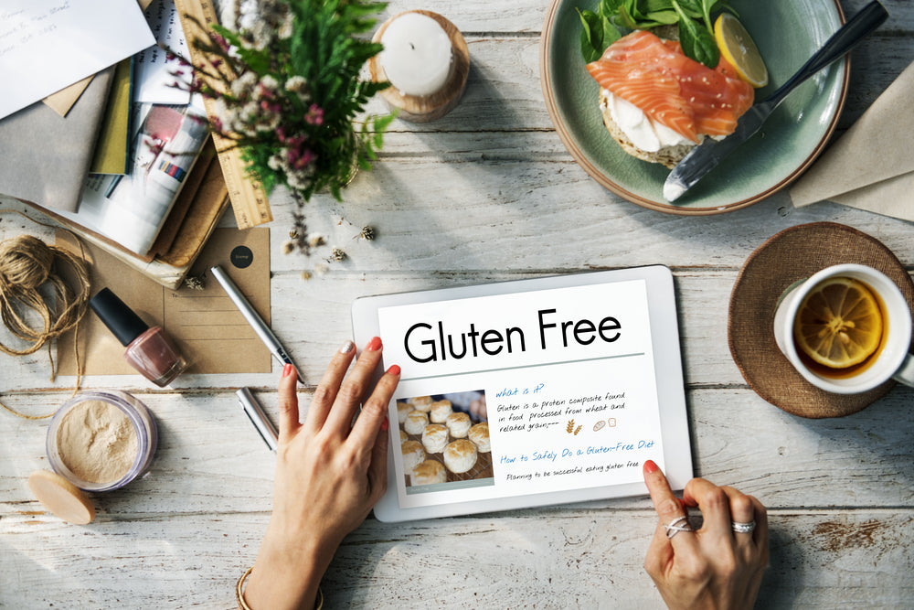 A hand holds a tablet displaying "Gluten Free" while surrounded by a cozy workspace with papers, a plate of salmon on a bagel, a cup of tea, and decorative flowers.