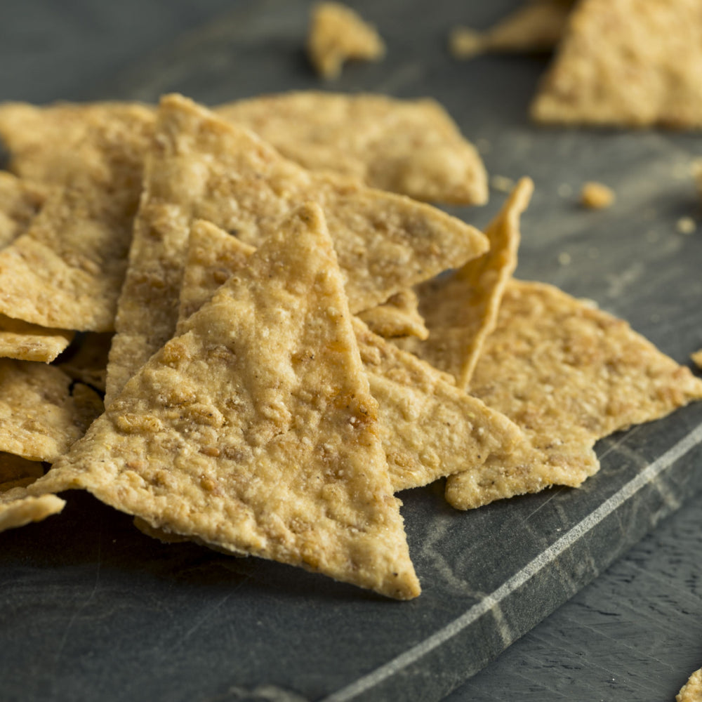 A close-up of a pile of golden-brown tortilla chips scattered on a dark stone surface, showcasing their triangular shapes and crispy texture.