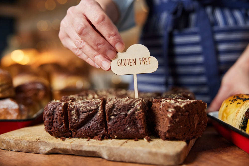 A hand places a wooden sign reading "GLUTEN FREE" into a freshly sliced brownie on a wooden board, with various baked goods blurred in the background.