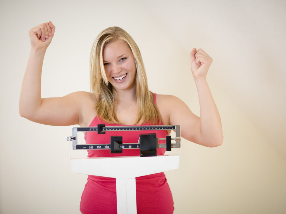 A smiling woman in a pink tank top raises her arms in celebration while standing on a scale, indicating a positive outcome in a bright, indoor setting.