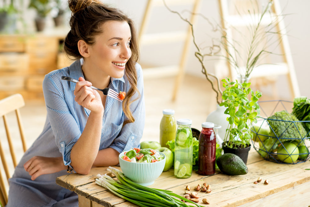 A smiling woman in a blue striped shirt sits at a wooden table, holding a fork with a piece of salad, surrounded by fresh vegetables and colorful juice bottles in a bright, airy setting.