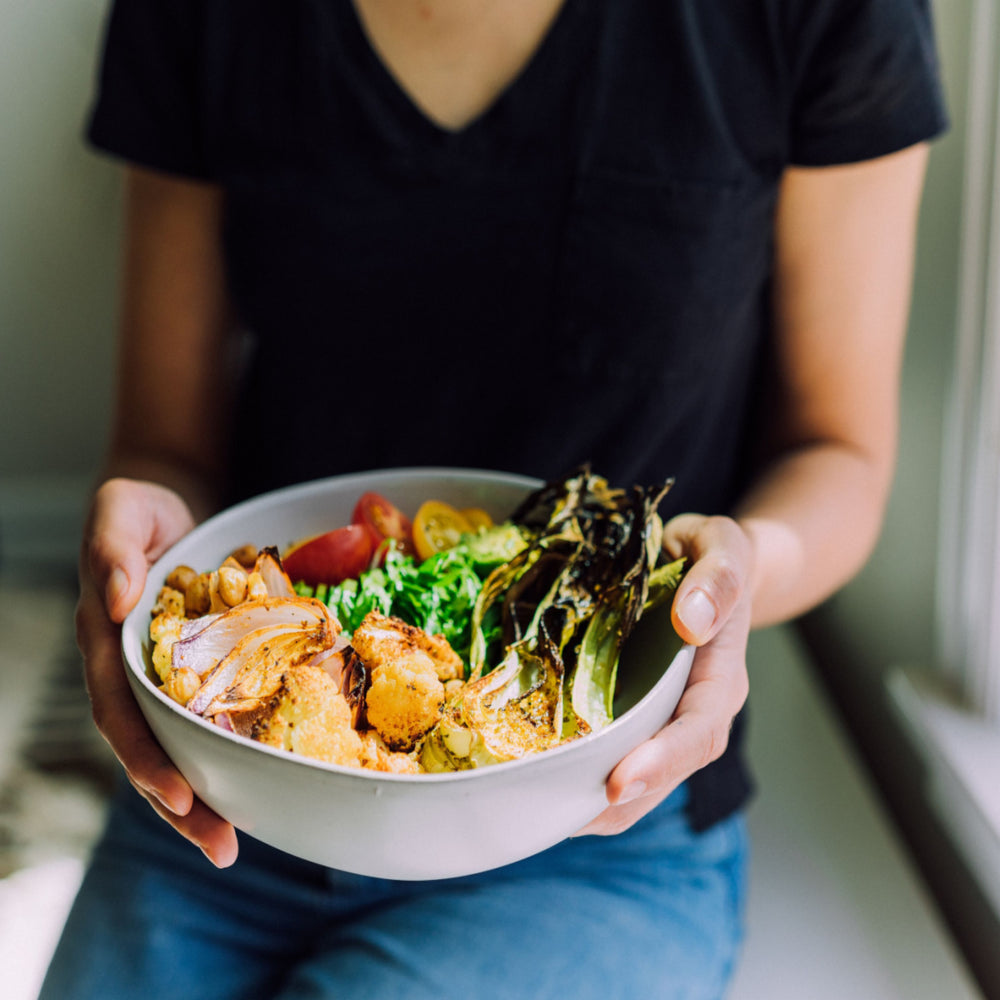 A person wearing a black shirt holds a bowl filled with roasted vegetables, chickpeas, and salad greens while sitting by a window.