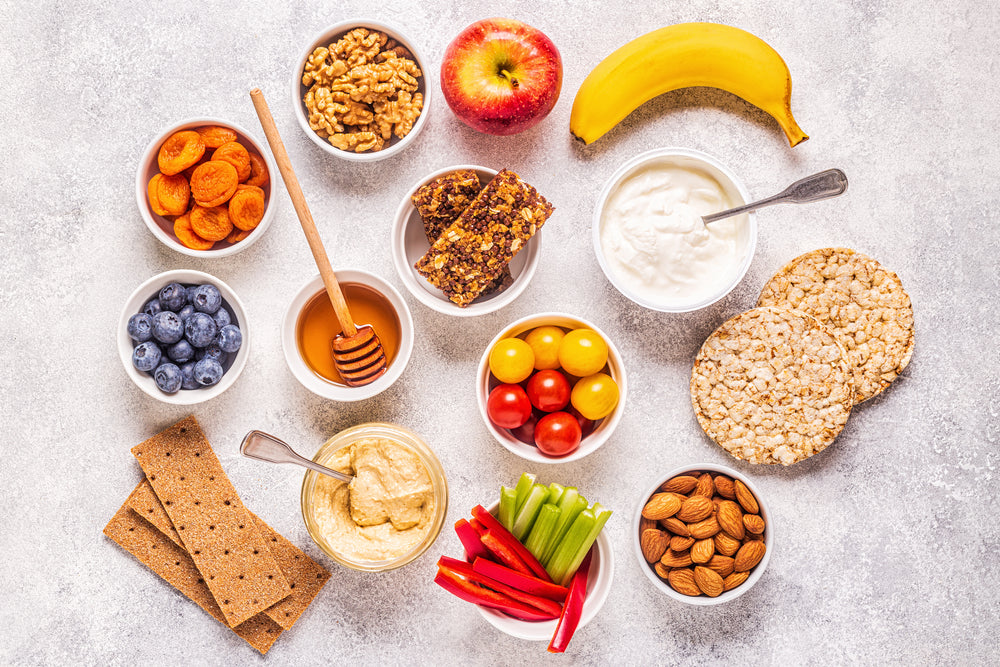 A variety of healthy snacks is arranged on a light surface, including fruits, nuts, yogurt, granola bars, hummus, and colorful vegetables in bowls, showcasing a vibrant and nutritious spread.