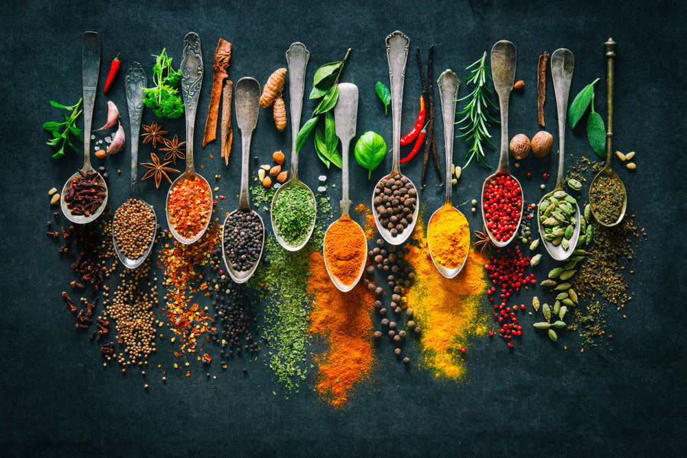 A variety of colorful spices and herbs are arranged in silver spoons on a dark surface, showcasing vibrant colors like red, yellow, green, and brown, highlighting their diverse textures and forms.