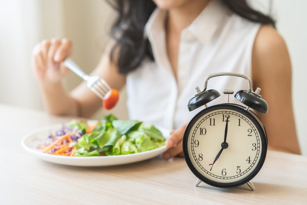 A woman in a white shirt is eating a salad with a fork while a black alarm clock sits on the table, indicating the time as she enjoys her meal.
