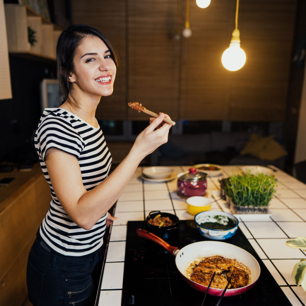 A woman with long dark hair smiles while holding a wooden spoon with food, standing in a modern kitchen filled with various dishes on a tiled countertop under warm lighting.