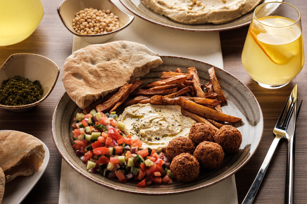 A plate features hummus, falafel balls, French fries, and a fresh salad with tomatoes and cucumbers, accompanied by pita bread and a glass of yellow drink, set on a wooden table.