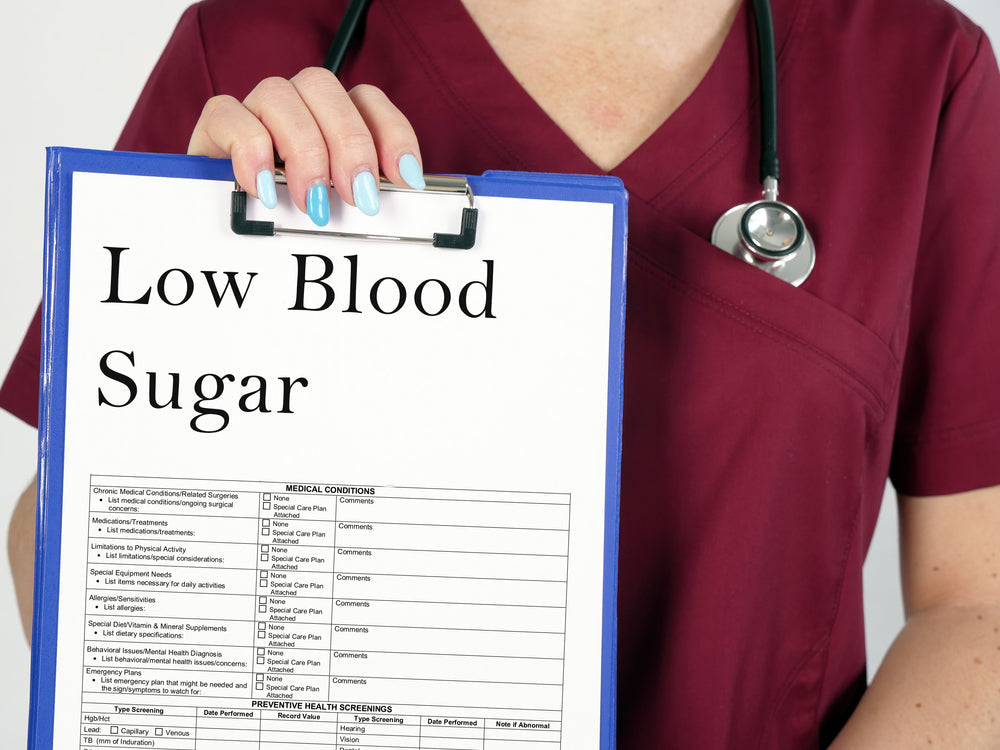A healthcare professional in a burgundy scrubs holds a clipboard labeled "Low Blood Sugar," displaying a list of medical conditions and preventive health screenings.