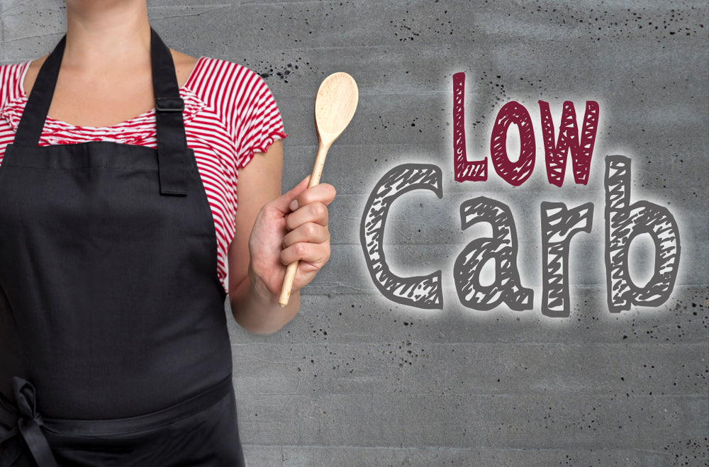 A person in a black apron holds a wooden spoon and stands in front of a gray wall featuring the words "Low Carb," emphasizing the theme of a low-carb diet for weight loss and health improvement.
