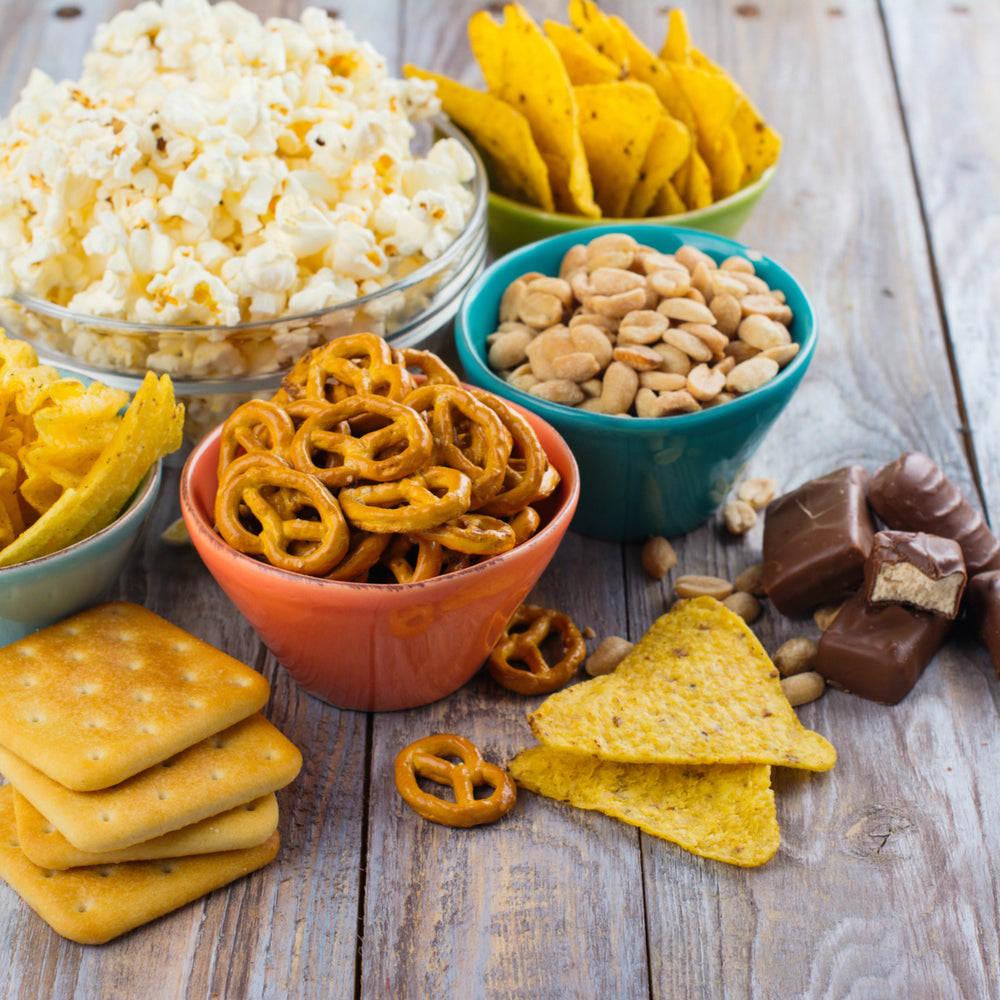 A variety of snacks is displayed on a wooden surface, including popcorn, pretzels, nachos, peanuts, chocolate bars, and crackers in colorful bowls and arranged attractively.