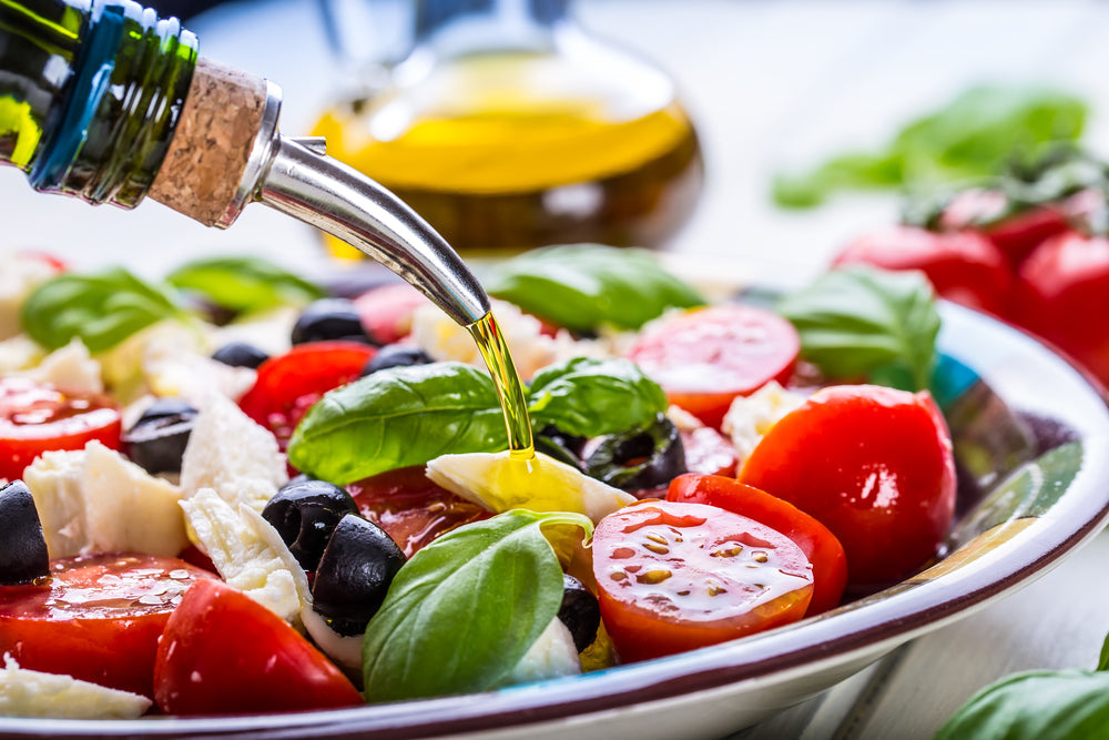 A bottle of olive oil is pouring over a fresh salad with sliced tomatoes, black olives, and basil leaves, set against a blurred background of additional ingredients and a glass bottle.