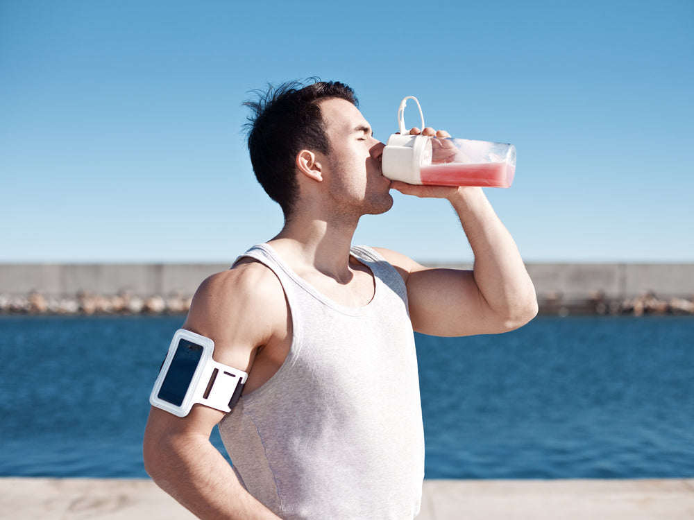 A man in a sleeveless shirt drinks from a clear shaker bottle while standing by a body of water, with a blue sky and a distant pier visible in the background.
