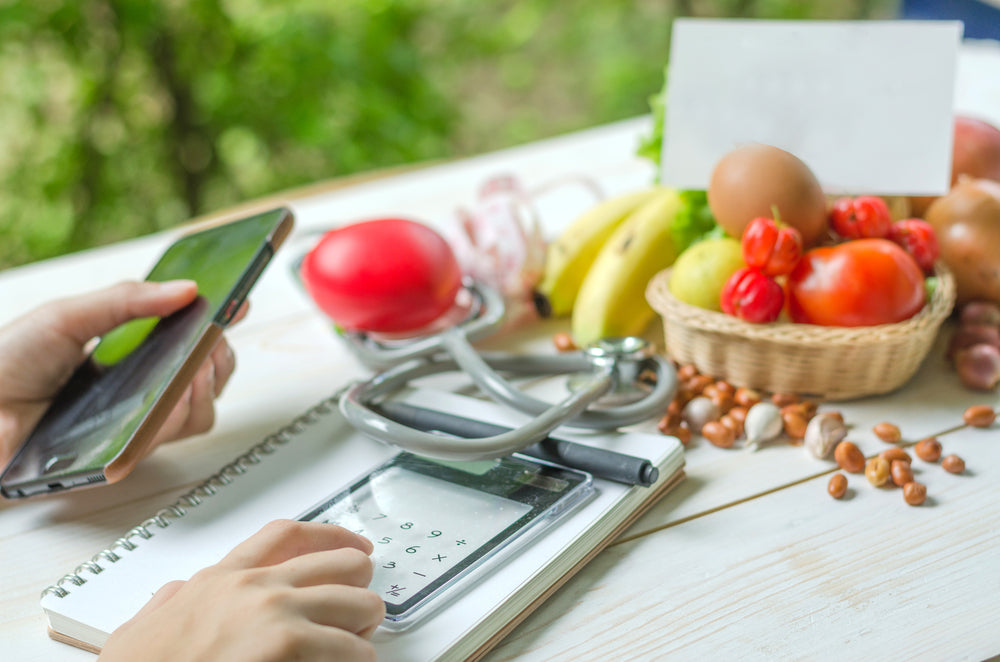 A person uses a smartphone and calculator while calculating net carbs, surrounded by fresh fruits, vegetables, and a stethoscope on a wooden table in a natural setting.