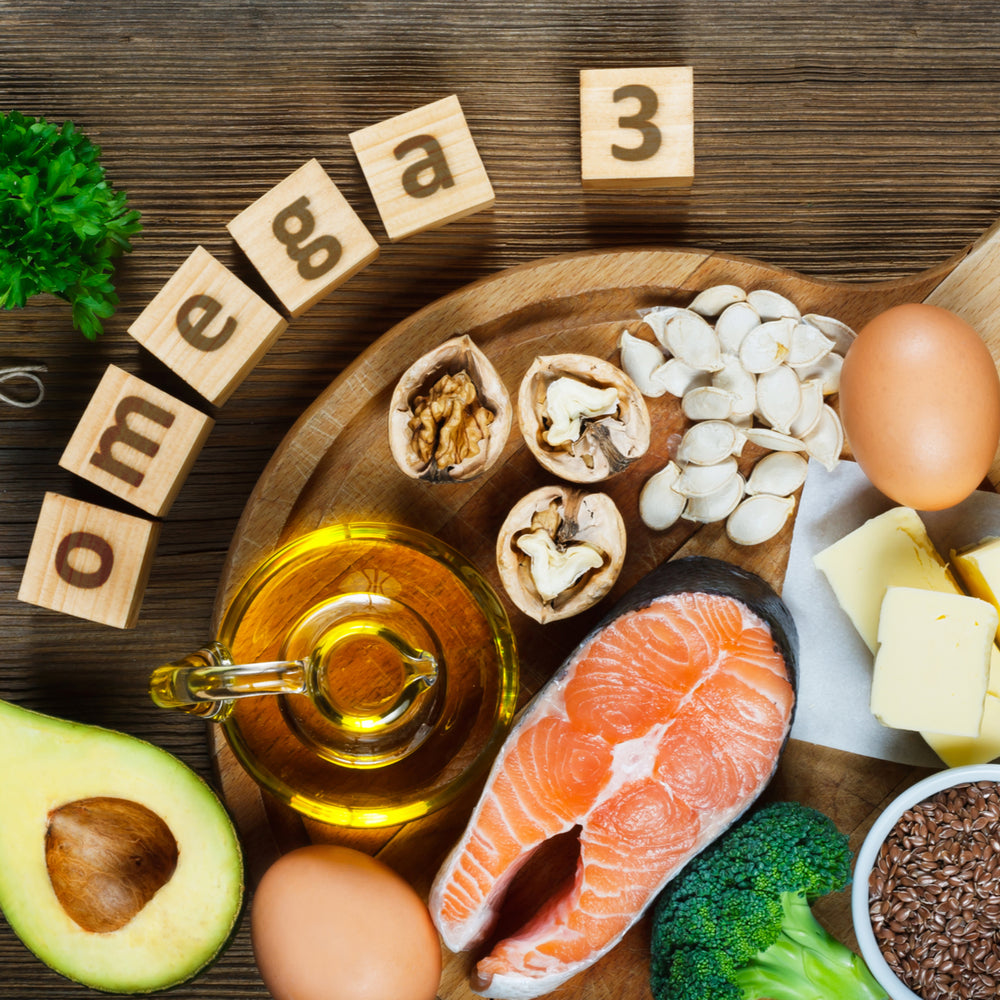 A wooden platter displays various foods rich in omega-3, including salmon, walnuts, flaxseeds, eggs, broccoli, and olive oil, with wooden blocks spelling "omega 3" nearby.