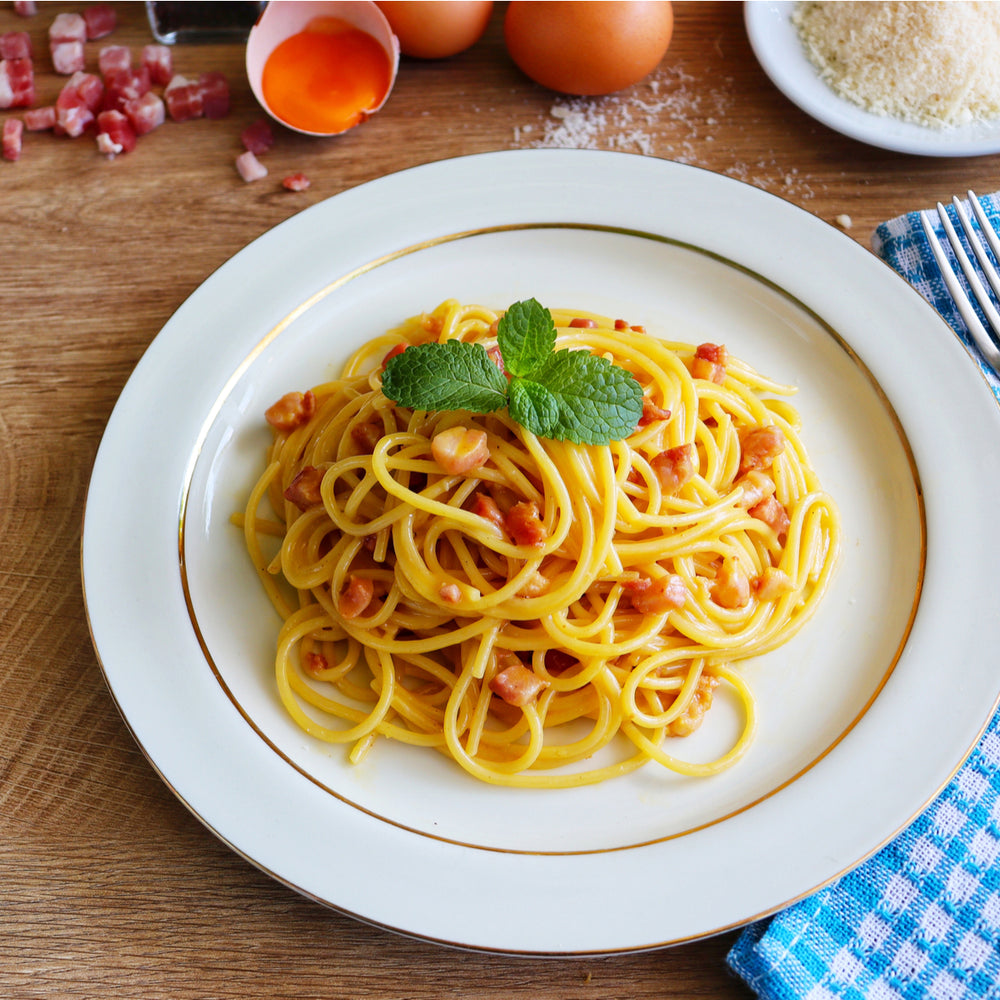 A plate of spaghetti is topped with pieces of bacon and a sprig of mint, surrounded by eggs, scattered bacon bits, grated cheese, and a blue checkered cloth on a wooden table.