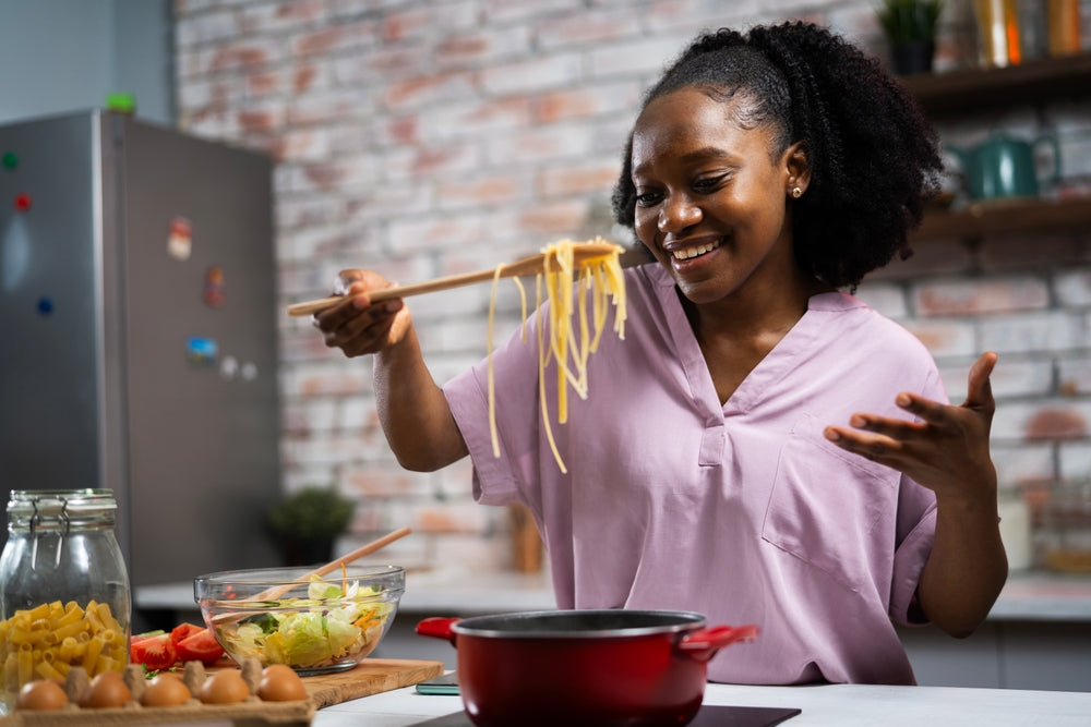 A woman in a pink shirt smiles while holding cooked spaghetti with chopsticks over a pot, surrounded by fresh vegetables and pasta in a modern kitchen with a brick wall.