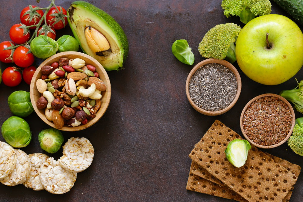 A variety of healthy foods is arranged on a dark surface, including a bowl of mixed nuts, fresh vegetables like Brussels sprouts and broccoli, an avocado, and green apple, alongside rice cakes and seeds.