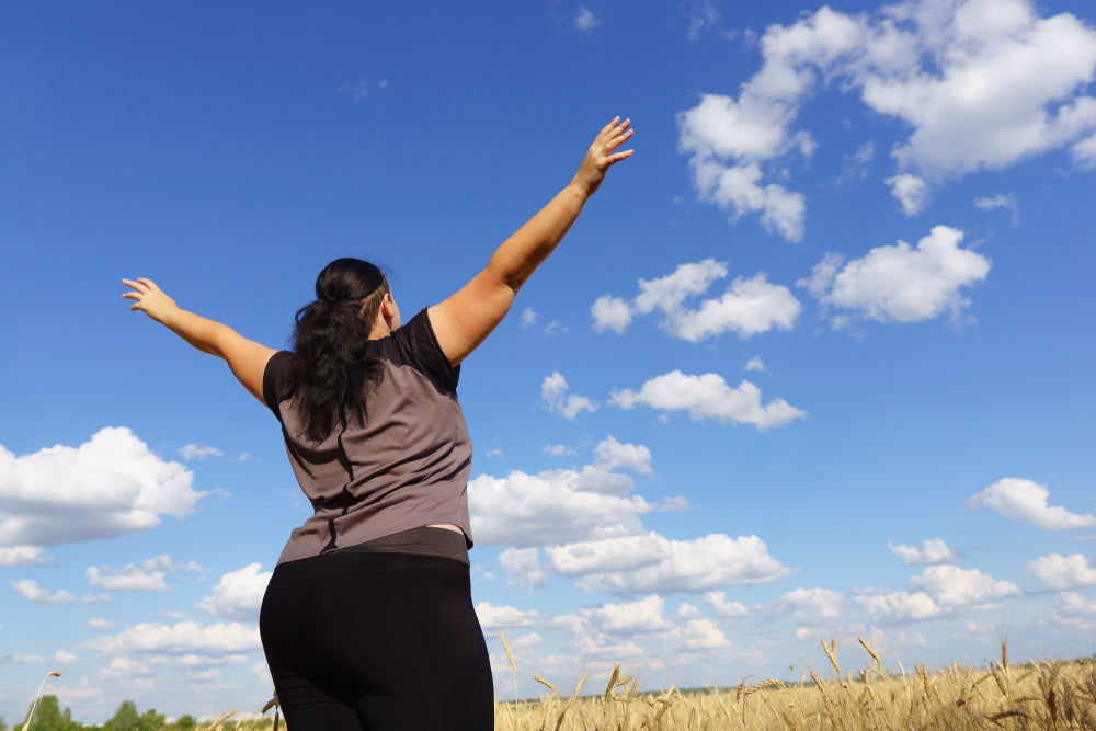 A person stands in a field with arms raised towards a bright blue sky, expressing joy and positivity, symbolizing a mindset shift for successful weight loss.