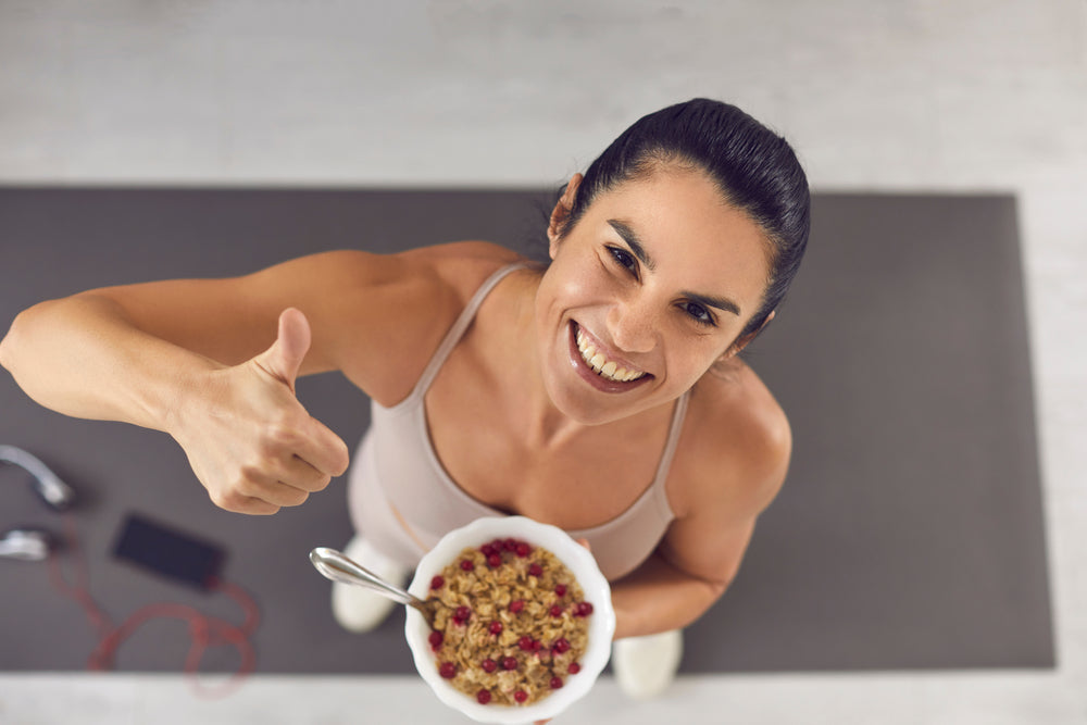 A smiling woman in athletic wear holds a bowl of oatmeal while giving a thumbs-up, standing on a yoga mat in a bright indoor setting, suggesting a positive pre-workout meal choice.