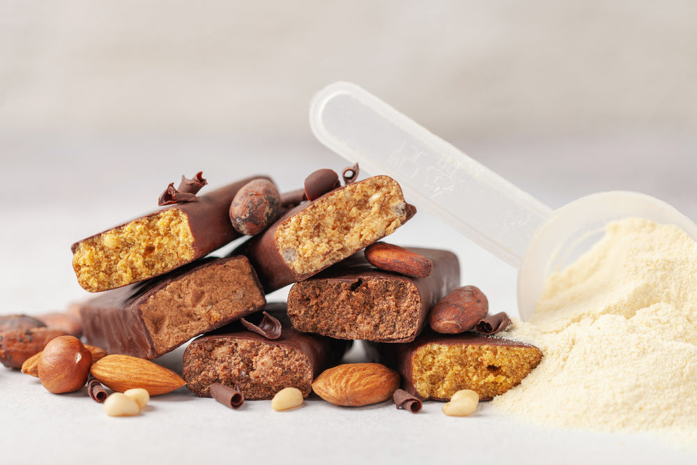 A stack of protein bars with different textures and colors sits next to a scoop and a pile of protein powder, surrounded by various nuts and chocolate shavings on a light background.