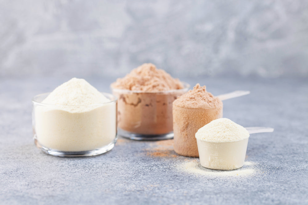 Four containers of protein powder in various flavors, including vanilla and chocolate, are arranged on a gray surface, showcasing different amounts and textures of the powders.